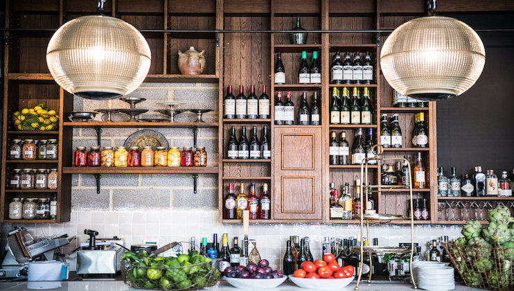 A well-organized kitchen shelf and counter space with various bottles of wine, jars of preserved foods, and fresh fruits and vegetables under two large, spherical, pendant lights. The background features a wooden pantry shelf with assorted items.