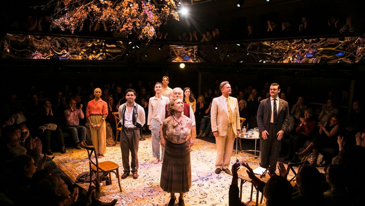 A theatrical cast stands on stage, looking up and holding hands, taking a bow after their performance. The stage is decorated with autumn leaves and rustic furniture. The audience surrounds them, clapping enthusiastically.