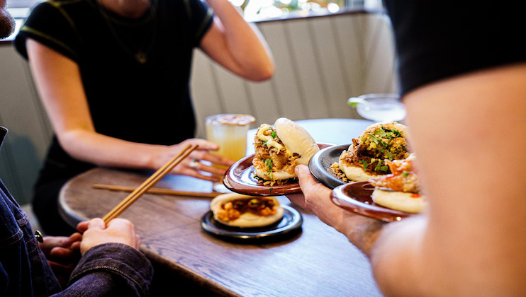 Two people are sitting at a table in a restaurant. One person holds chopsticks while the other is served bao buns with various toppings on small plates. The atmosphere is casual and the setting has a cozy vibe. Drinks are also visible on the table.