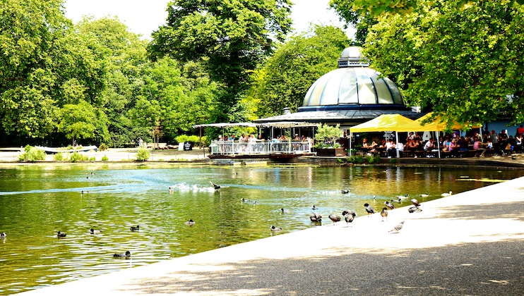 Scenic view of a park with a large pond. Birds, including ducks and swans, are swimming in the water and resting near the edge. People are seated under a pavilion with yellow awnings on the far side of the pond, surrounded by lush green trees.