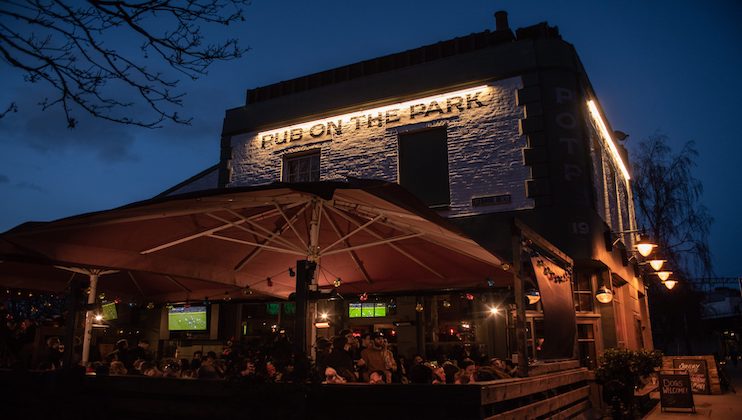 A bustling evening scene at Pub On The Park. Outdoor seating under large umbrellas is filled with people, warmly lit by string lights. The brick building features a neon sign, while trees and a dark blue sky create a cozy atmosphere.