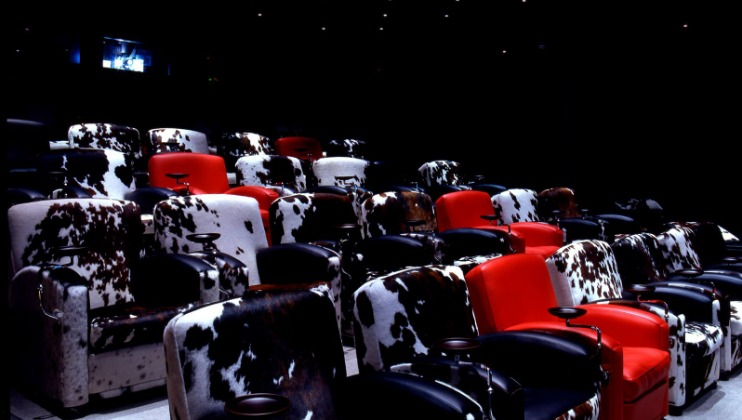 A dimly-lit movie theater with unique, plush, cow-print and red reclining seats. The chairs alternate between black and white cow prints, and solid red upholstery. Each seat is equipped with a cup holder. The ambiance is cozy and intimate.