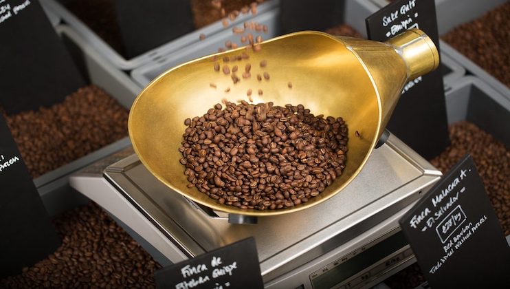 A large brass scoop is filled with roasted coffee beans, some of which are spilling out. The scoop is resting on an electronic scale. Several black signs with text in chalk are in the background, likely indicating different coffee bean varieties.