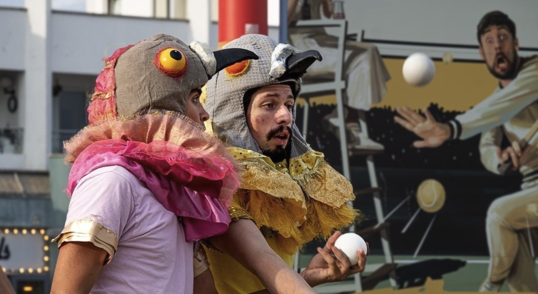 Two street performers, wearing whimsical bird costumes with oversized eyes and colorful frills, juggle white balls in front of a mural depicting tennis players in action. The background consists of urban buildings and onlookers watching the performance.