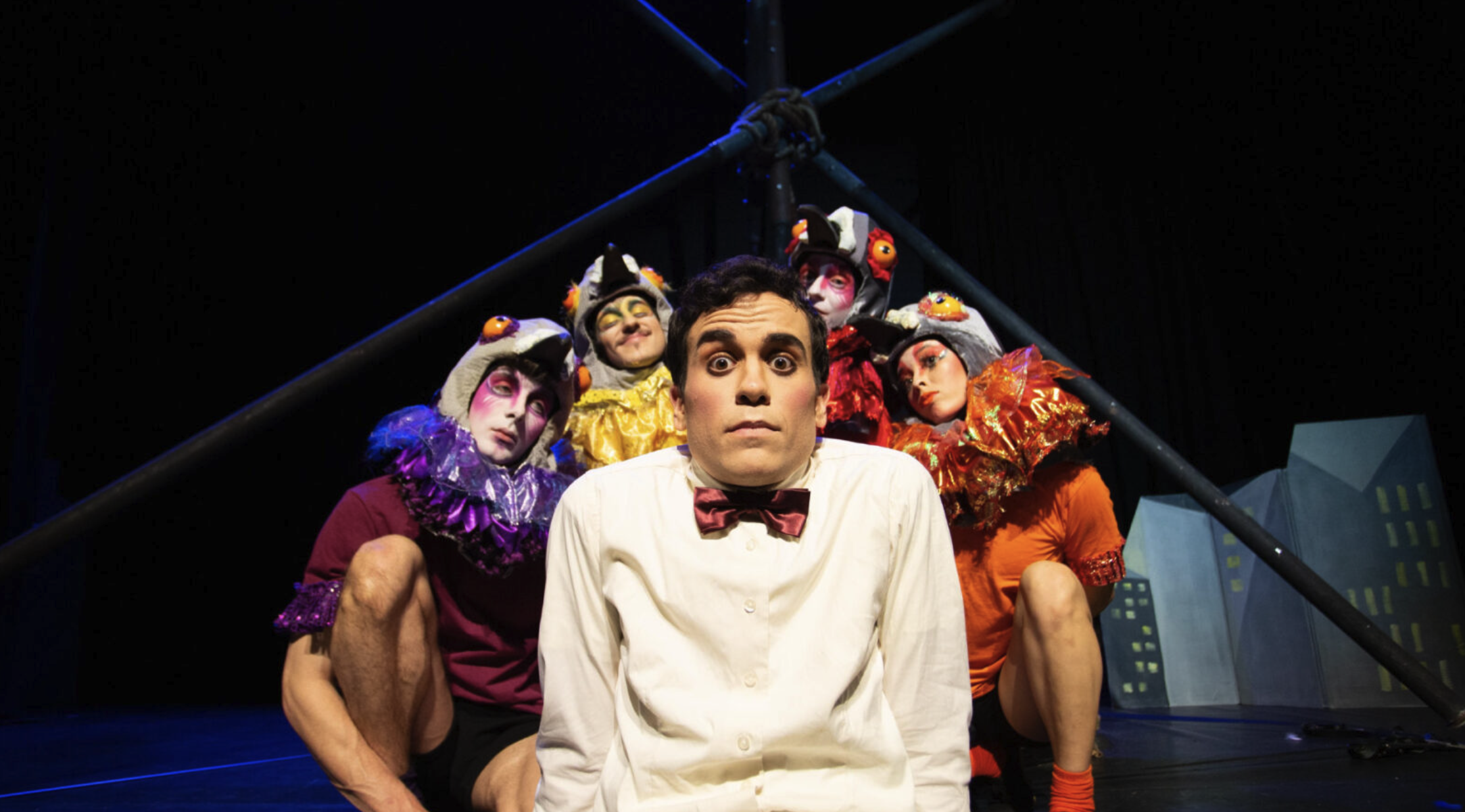 A man in a white shirt with a red bow tie sits in the foreground, looking surprised. Behind him, four people wearing colorful bird costumes, complete with detailed masks and feathered collars, strike dramatic poses on a dimly lit stage with a dark, industrial backdrop.