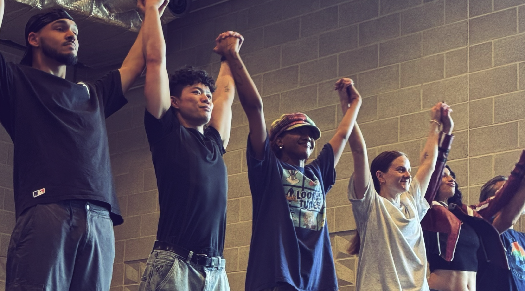 Five people stand in a line in a room with grey cinder block walls, raising their hands together in celebration. They appear to be in casual attire and display expressions of joy and unity.