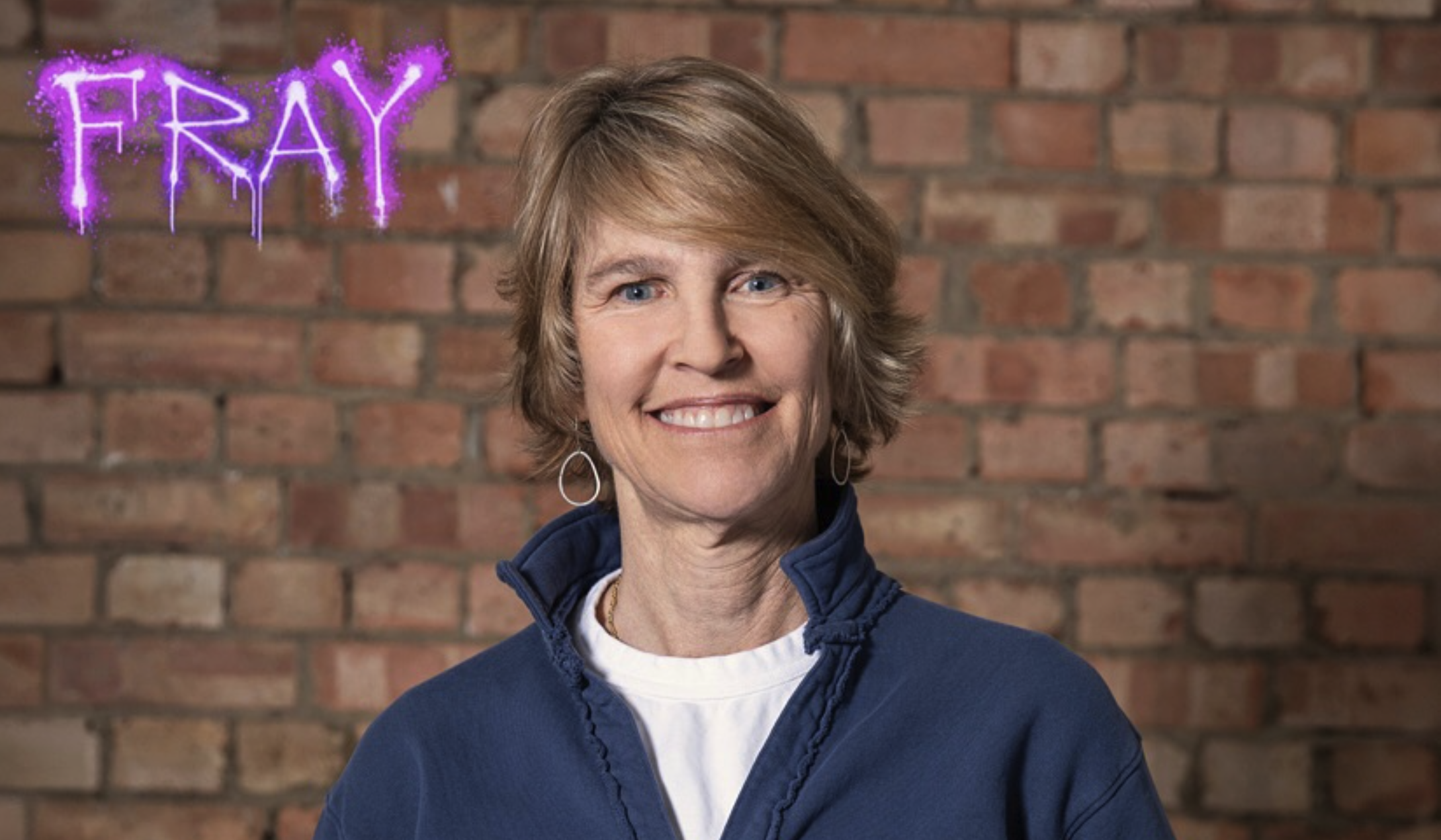 A smiling person with short hair is posing in front of a brick wall. They are wearing a blue jacket over a white shirt. The word FRAY is written in purple, glowing text in the top left corner of the image.