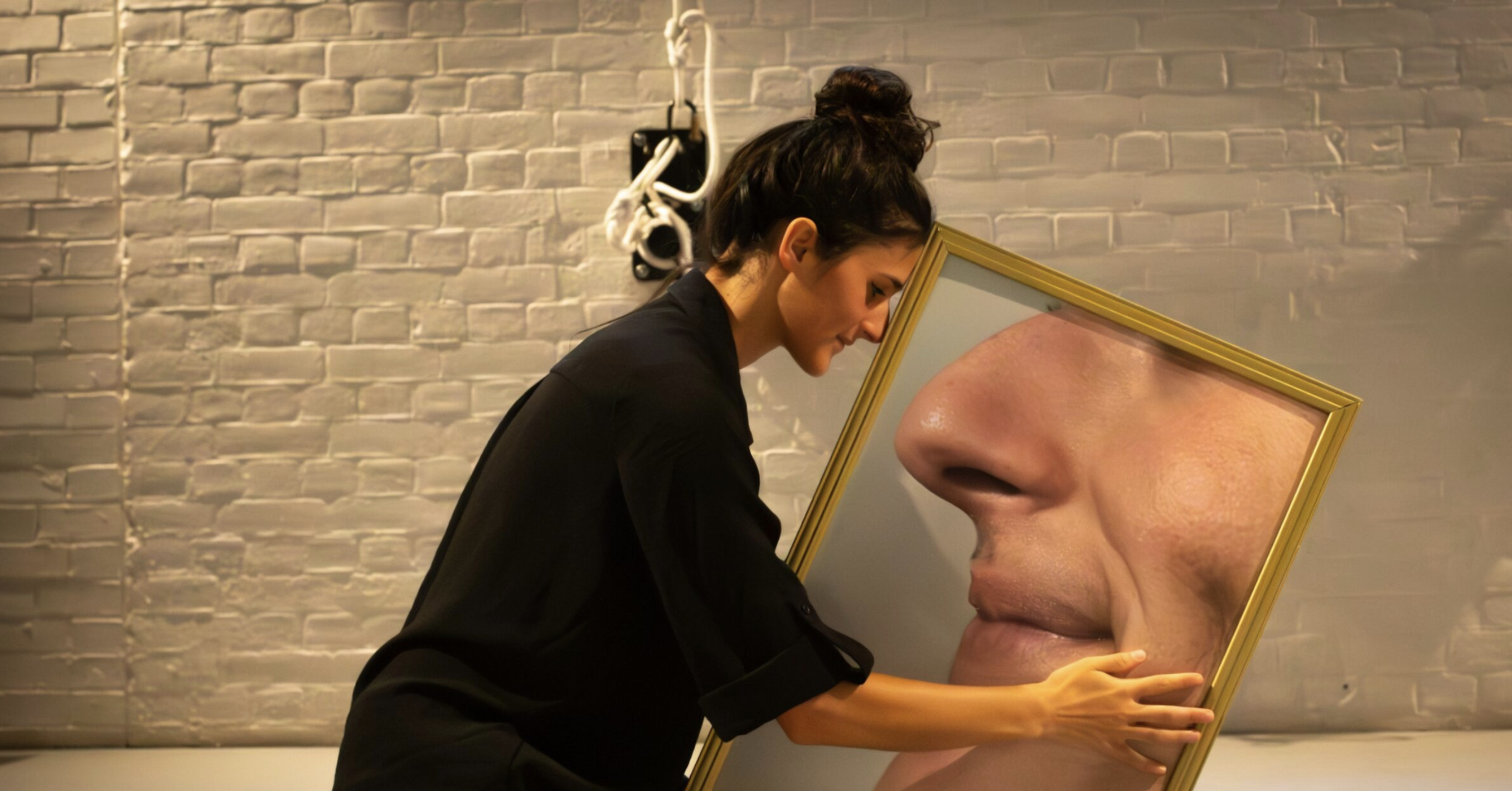 A person dressed in black is leaning towards and embracing a large mirror that shows a close-up reflection of their nose and mouth. The background consists of a white brick wall with a white cable and fixture hanging from it.