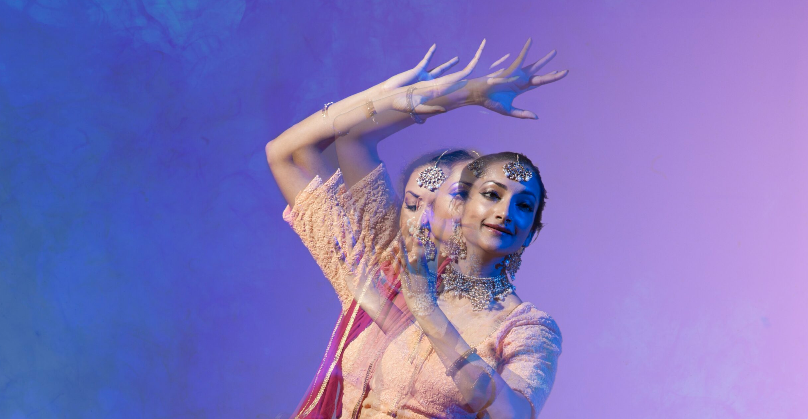 A woman wearing traditional Indian attire, including jewelry and hair accessories, performs a classical dance. Her movements are captured in a double exposure effect against a purple gradient background, emphasizing the grace and fluidity of her dance.