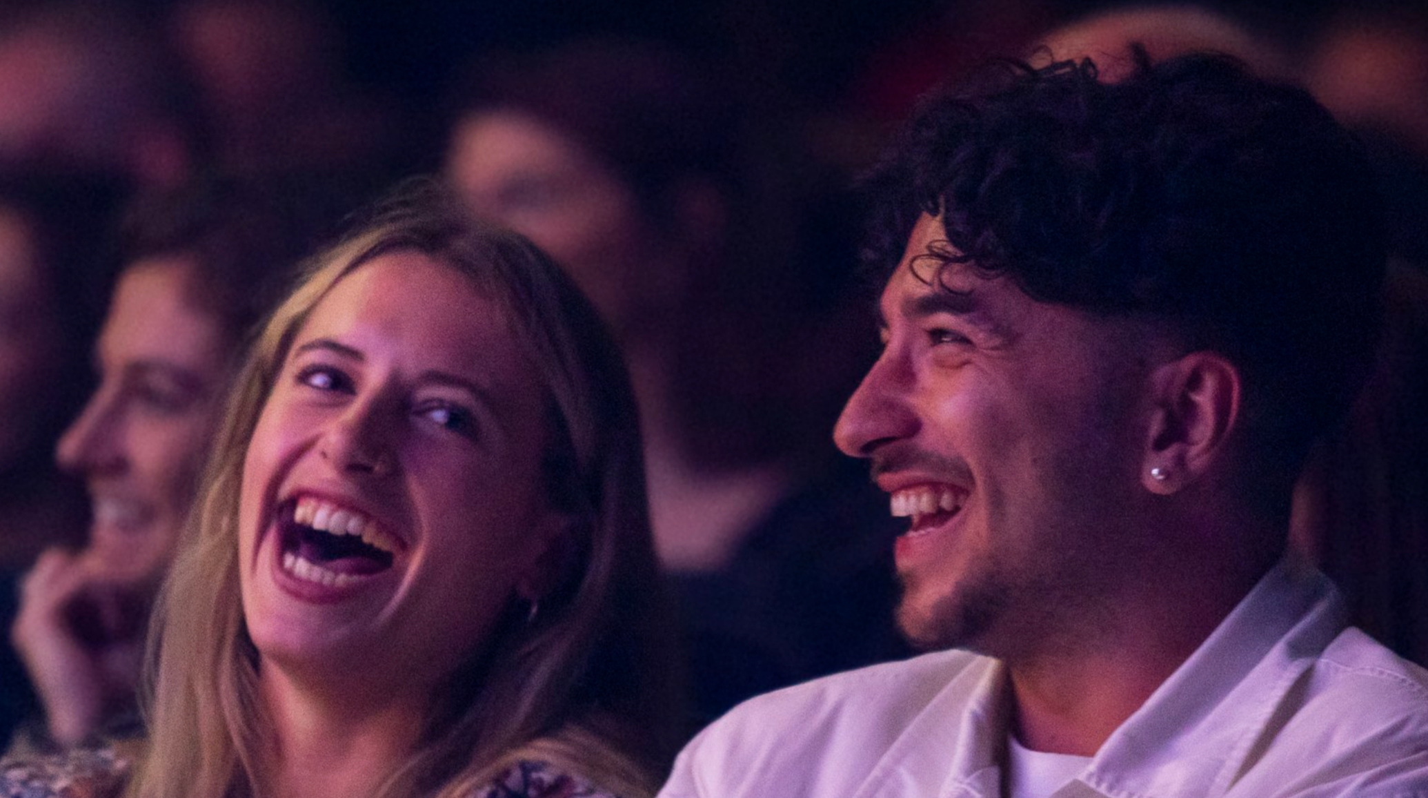 A woman and a man, both with light skin, sit closely together in a dimly lit audience. They are laughing and looking at each other with joy. The background is blurred, showing other audience members who appear to be enjoying the event as well.