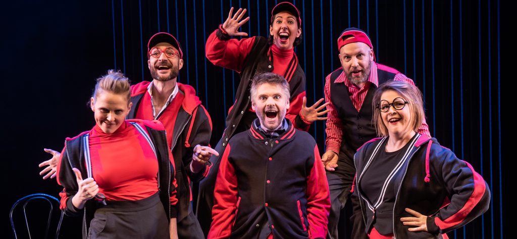 A group of six people, wearing matching black and red outfits, pose with animated expressions on stage. Some are smiling broadly, while others make playful gestures. A dark background and stage lights highlight their energetic and joyful demeanor.