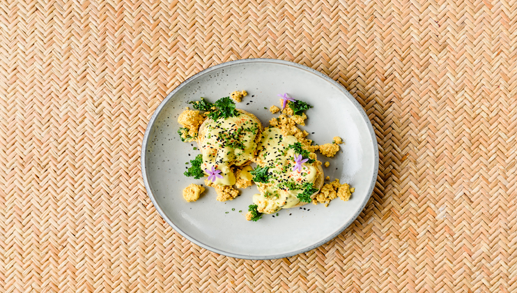 A white plate with an artistic arrangement of food, featuring two scoops of a creamy, yellow substance garnished with green herbs and small purple flowers. Crumbled bits of a light-colored topping are sprinkled around the plate. The background is a woven, tan mat.