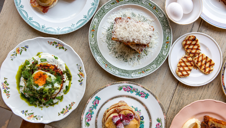 A wooden table set with various breakfast dishes, including avocado toast topped with an egg and green garnish, pancakes with fruit, grated cheese on toast, grilled heart-shaped pastries, boiled eggs, and a sandwich. The dishes are served on decorative plates.