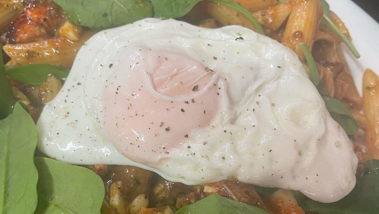 A close-up of a dish featuring penne pasta coated in a flavorful sauce, mixed with vegetables and topped with a perfectly cooked, slightly runny fried egg. Fresh green leafy vegetables garnish the dish, adding a touch of color and freshness.