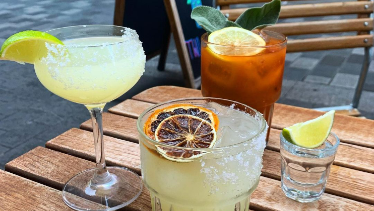 Close-up of a wooden table with three cocktails. A margarita with a salted rim and lime garnish, a cocktail with an orange slice and leafy garnish, and a drink with a dried citrus slice on top. A shot glass with a lime wedge is next to the foreground cocktail.