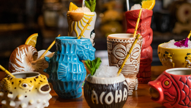 A colorful assortment of tiki mugs filled with cocktails, each uniquely designed. Mugs resemble various tropical and sea creatures with intricate patterns. Some are garnished with fruit slices, mint leaves, or flowers, and all have straws for drinking.