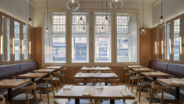 A modern restaurant interior with wooden tables and chairs neatly arranged. Large windows with geometric glass patterns allow natural light to fill the space. Hanging light fixtures add an elegant touch, and simple table settings are ready for guests.