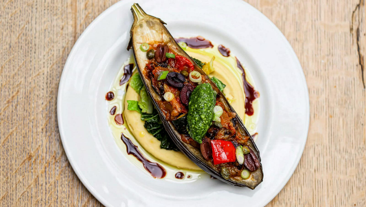 A beautifully plated dish featuring a grilled eggplant boat stuffed with a vibrant mix of vegetables and garnished with green herbs. The eggplant sits atop a swirl of creamy sauce on a white plate, drizzled with balsamic reduction and surrounded by leafy greens.