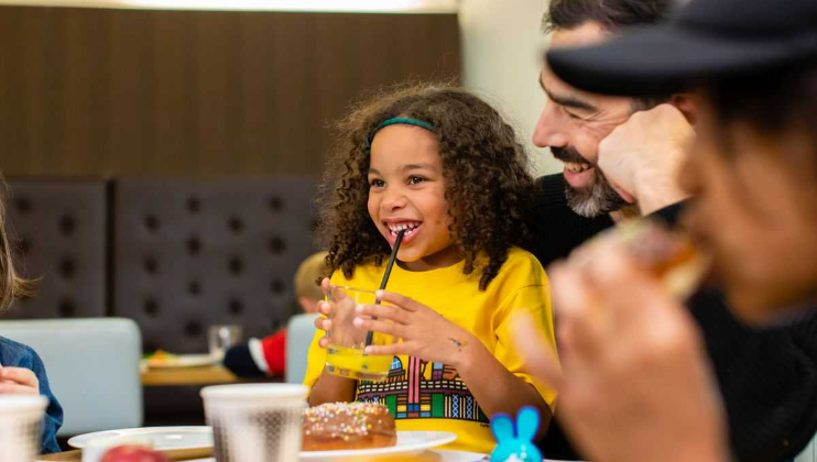 A young girl with curly hair, wearing a yellow shirt, smiles while sipping a drink through a straw at a dining table. A man next to her with a beard is looking at her, smiling. Other people are partially visible in the background. The table has food and drinks.