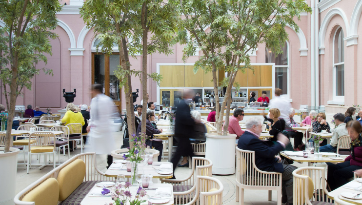 A lively indoor café with a high ceiling and pink walls. Patrons sit at tables decorated with small plants, engaged in conversation. Waitstaff in white attire move swiftly between tables. Tall potted trees are scattered throughout the seating area.