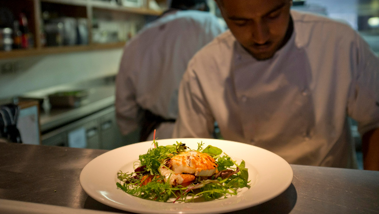 A chef in a white uniform focuses intently on preparing a gourmet dish. The plate in the foreground features a beautifully presented meal with greens and a central main dish, likely seafood, garnished with herbs, sauce, and microgreens, in a professional kitchen setting.