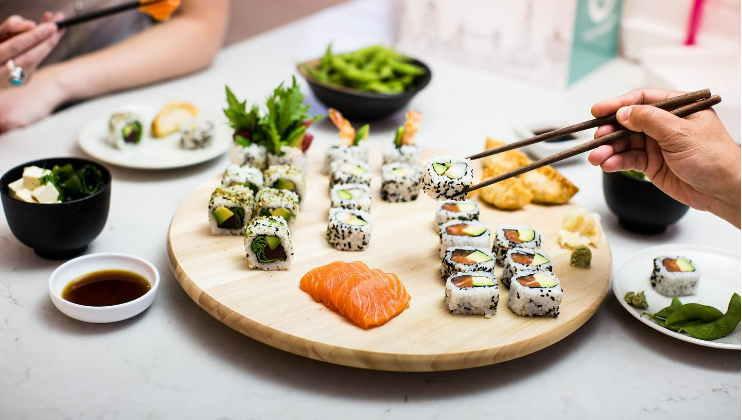 A wooden platter is filled with various sushi rolls and slices of sashimi, surrounded by dipping sauces and garnishes on a white table. Two people are eating with chopsticks, while bowls of edamame, miso soup, and other dishes are also visible in the background.