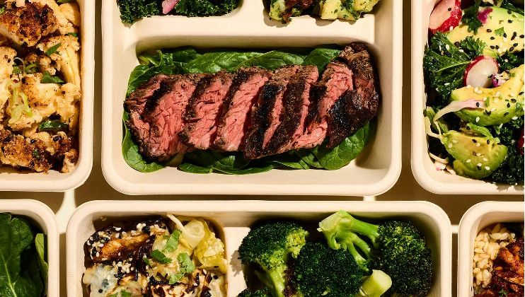 An assortment of meal containers featuring various foods, including grilled steak slices on a bed of greens, broccoli florets, a salad with avocado and radishes, roasted cauliflower, and other mixed vegetables. The meals are neatly arranged in a grid-like pattern.