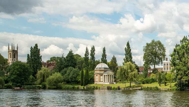 Riverside scene with a domed building, surrounded by lush greenery and tall trees, and a church tower visible in the background. The sky is partly cloudy, and the calm water reflects the landscape.