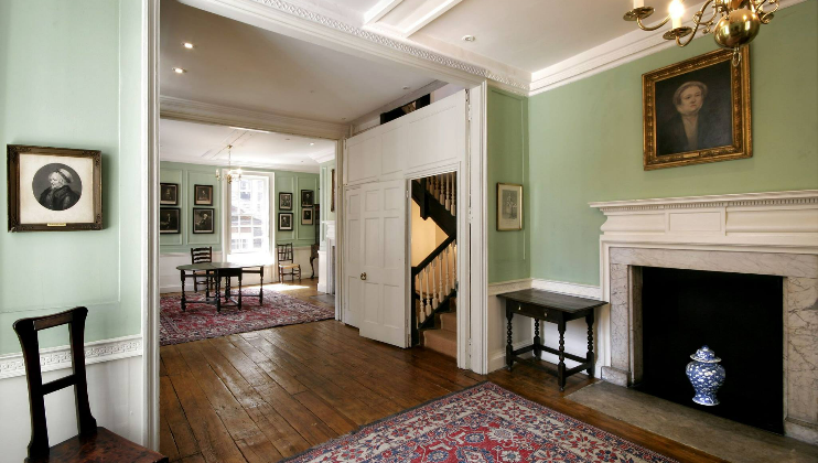 A spacious, well-lit room with wooden floors and green walls, featuring framed portraits, white-paneled doors, a fireplace with a marble mantle, and an ornate vase. The room has antique furniture, including chairs and tables, and patterned rugs. A staircase is visible in the back.