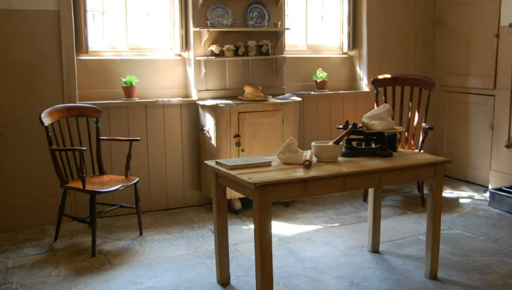 A rustic kitchen with wooden chairs, a small wooden table, and a shelf with blue and white plates. The table has a mortar and pestle, cloth, and a scale. Potted plants decorate the windowsill, with sunlight streaming through the windows.