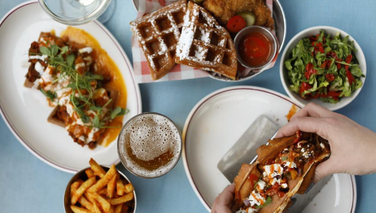 Top-down view of a table with assorted dishes: loaded waffles, fried chicken with waffles, a bowl of salad, a serving of fries, a glass of beer, and another beverage. A person is holding a taco, ready to eat. Plates and utensils are on a light blue tablecloth.