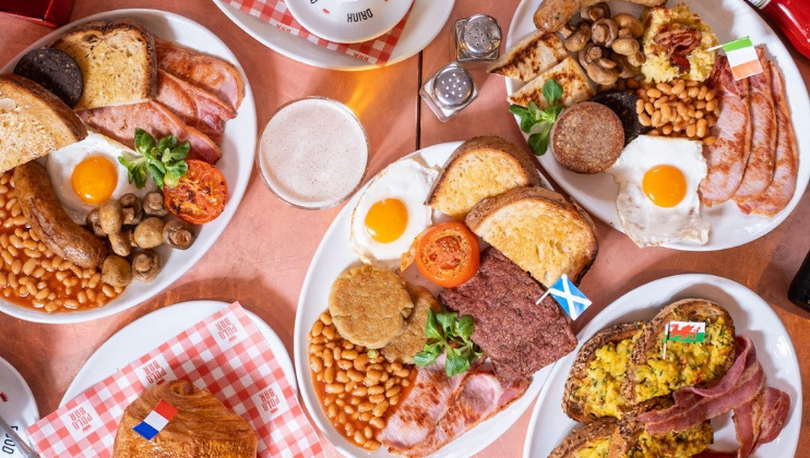 Five plates of breakfast foods, each featuring different items: fried eggs, bacon, baked beans, toast, sausages, mushrooms, tomatoes, and different flags. A croissant on a checkered napkin is in the bottom left. Salt and pepper shakers are placed in the center.