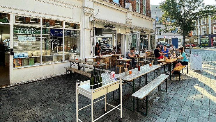 Outdoor seating area of a cafe with wooden tables and benches. A few patrons are seated, engaged in conversation and dining. The cafe is situated on a cobblestone street and has large windows and an open entrance. Greenery is visible in the background.