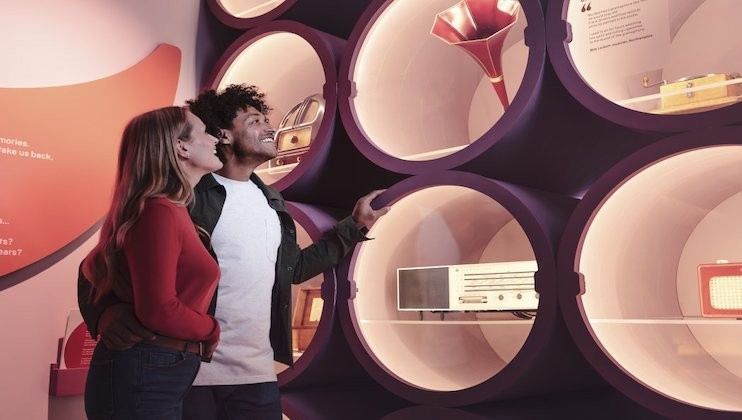 A couple stands in front of a display of vintage audio equipment in a museum. The items are showcased in round, illuminated compartments. They include gramophones, radios, and other antique devices. The couple looks interested and engaged.