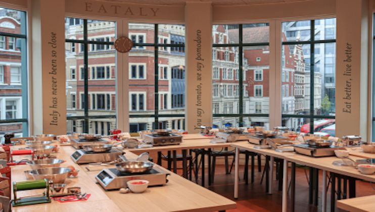 A classroom with large windows overlooking a city street is set up with rows of tables holding cooking equipment, such as burners, pots, and cutting boards. The walls display cooking-related quotes, and the seating is arranged to accommodate multiple students.