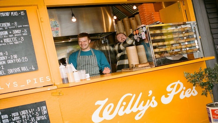 Two people smiling and serving at a bright yellow food stall named Willy's Pies. Pies and serving cups are visible on the counter. A chalkboard menu to the left lists various pie options and prices. The interior of the stall has warm, ambient lighting.