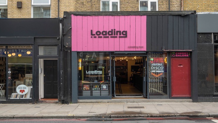 Street view of a building with three businesses. The central business has a pink facade and signage that reads Loading with Loading Bar underneath. To the right is a place advertising cocktails, disco, and late nights. To the left, there is a shop with books displayed.