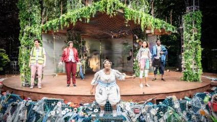 A theatre performance set outdoors features five performers on stage adorned with greenery and vines. The stage is surrounded by netting and assorted debris, suggesting an ecological theme. The actors are in varied, colorful costumes. Trees are visible in the background.