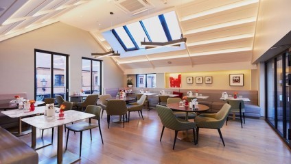 A modern café features a bright, airy interior with large skylights and windows. Tables are neatly arranged with green and brown chairs. Neutral tones dominate the decor, and framed art pieces hang on the walls, enhancing the comfortable ambiance.