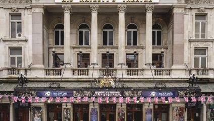 Façade of His Majesty's Theatre in London, displaying banners for Phantom of the Opera. The building features a grand entrance with tall columns and a balcony, adorned with British flags above the awning. The theatre's name is prominently displayed at the top.
