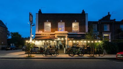 A cozy pub named Prince George is illuminated with string lights in the evening. Bicycles are parked outside the entrance. The pub has large windows on the first floor, and a sign at the entrance advertises Cellar Cooled and Real Ale.