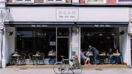 A street view of BEAM café, deli, and bistro with outdoor seating. People are dining at tables under a black awning. A green bicycle is parked in front of the establishment on the sidewalk. The building facade is light with large windows.