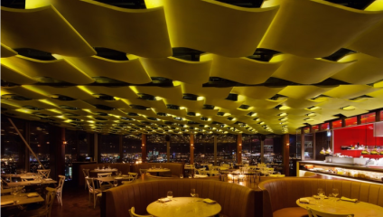 A modern, upscale restaurant with a yellow, wave-like ceiling design and panoramic city views at night. The dimly lit interior features round tables with elegant place settings, tufted booths, and a sleek, illuminated bar area.