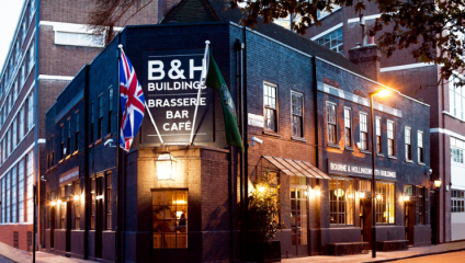 A dark brick building with a sign reading B&H Building Brasserie Bar Café on the corner, adorned with outdoor lights and a Union Jack flag. The building has large windows and awnings, with trees and adjacent structures visible in the background.