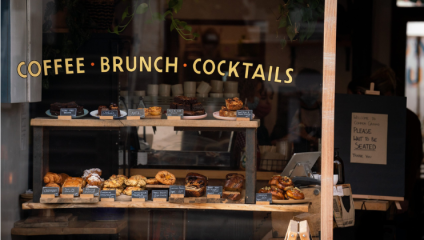 A cozy café window display featuring various baked goods, including pastries, croissants, and cakes. Above the display, a sign reads COFFEE • BRUNCH • COCKTAILS. A chalkboard sign inside the café reads, Welcome to Corner Café, please wait to be seated, thank you.