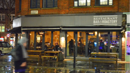 A cozy restaurant with a sign reading Restaurant Bar & Roastery on a rainy evening, located on Exmouth Market. The warm interior lighting contrasts with the wet street outside. People inside are dining, while a few individuals are seated and walking outside.