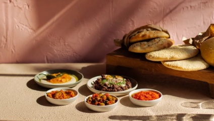 A selection of dishes is displayed on a beige table against a pink background. Small white bowls contain various colorful foods, and a wooden tray holds different types of bread, including flatbreads and rolls. The scene is bathed in warm, natural light.