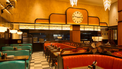 A retro-style diner with teal and red leather booths. The room features a large clock on the wall, an art-deco design with warm yellow and brown colors, and hanging pendant lights. Tables are set with utensils and menus, giving a classic, vintage atmosphere.