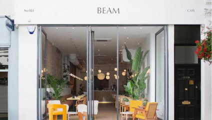 Front view of the café BEAM with a modern aesthetic. Features large glass windows revealing a bright interior with yellow and wooden chairs, tables, and leafy plants. Pendant lights hang from the ceiling, and No 103 is marked on the white facade.