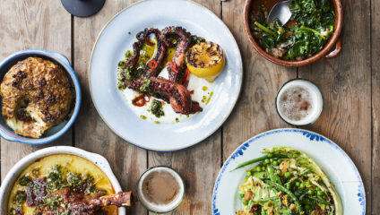 An overhead view of a rustic wooden table set with various dishes. There's grilled octopus with a lemon half, a dish of roasted cauliflower, a bowl of leafy greens, a meaty casserole, and a fresh green salad. Two glasses of beer are also on the table.